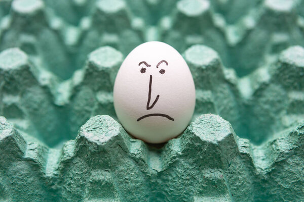 Крупный план белого куриного яйца с сердитым лицом, изолированным на зеленом фоне картонной коробки. Печаль, эмоции, выражение, грусть, одиночество.
