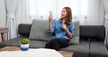 Evde akıllı telefonlu Asyalı genç bir kadının portresi. Çekici kız kanepede oturmuş, rahat bir yerde arkadaşlarıyla sohbet ediyor.