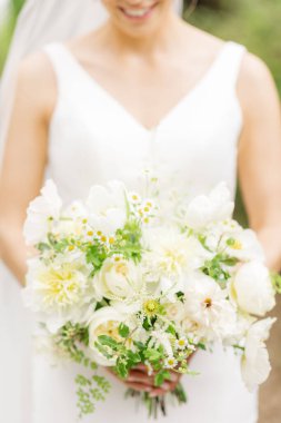 Beyaz bir elbise giymiş, elinde bir buket beyaz gül ve yeşil yaprak tutan bir gelinin dikey görüntüsü.