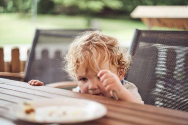 Sarışın, beyaz bir çocuk dışarıda bir masada oturmuş yemek yiyor.