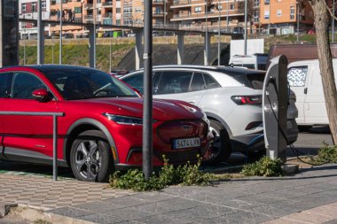 Two electric vehicles, a red Ford Mustang and a white Volkswagen, charging their batteries in a public parking lot, clean and renewable energy. clipart