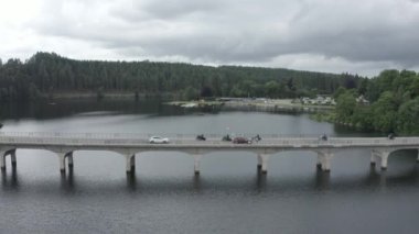 A high angle drone footage of motorcycles racing on a bridge road above a lake at a village