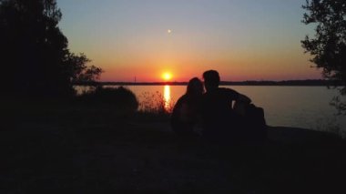 A young couple sitting at the coastline of the lake and looking at the beautiful sunset