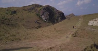 A drone footage of two hikers walking on a narrow path on the mountainous shore in Batanes, Philippines
