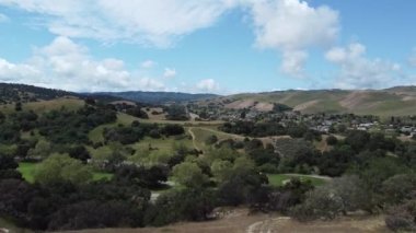 Mavi bulutlu gökyüzüne karşı ağaçlı ve tarlalı bir arazinin hava aracı görüntüsü.