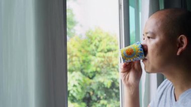 A Southeast Asian male drinking coffee and looking from a window in the early morning
