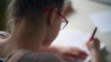 A closeup of a Caucasian female in glasses seen from behind, sitting at the table, taking notes