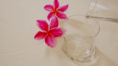 A closeup of water poured into an empty glass with pink frangipani flowers