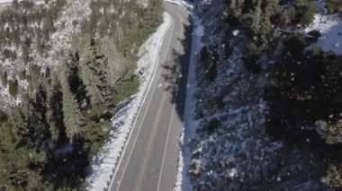 An aerial view of a two-line asphalt road passing through a forest covered in snow on a sunny day
