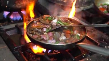 A close up of frying pan with meat and vegetables inside, in slow motion, on high heat in an industrial kitchen