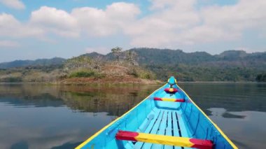 A bright blue and yellow wooden boat sailing across a lake surrounded by lush wilderness under a bright cloudy sky