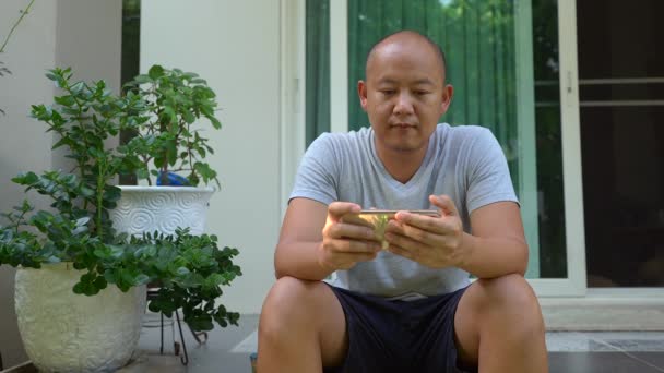 Bald Southeast Asian Man Playing His Phone While Sitting Patio — Stock videók