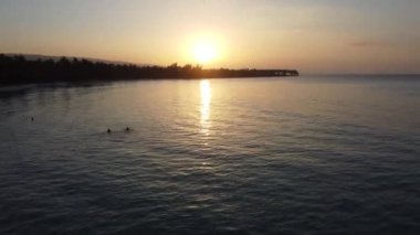 Kıyı şeridinin arka planında insanların olduğu güzel mavi bir deniz ve gün batımında gökyüzü renklendiriliyor.