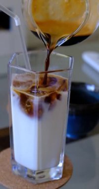 A vertical shot of coffee being poured into a glass of milk with ice