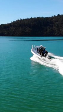 The vertical drone footage of the white boat taking a tour on a turquoise calm and clean ocean water  around the mountainous land, New Zealand