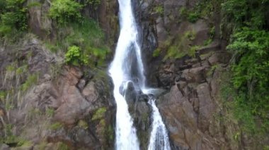 An aerial view of a beautiful mountain waterfall with green plants and trees growing around on a sunny day