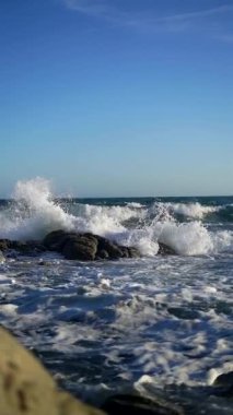 Dalgaların yavaş çekimde mavi gökyüzünün altında devasa taşlara çarpması.