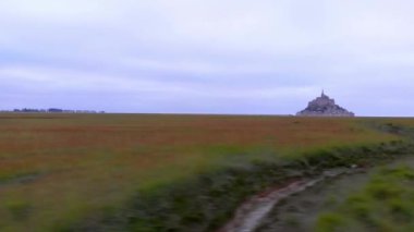 A scenic shot of a long green grass field and a stone hill in the background surrounded by water