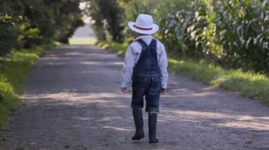 The footage of a little boy with a white cowboy hat walking along the rural road.