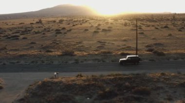 An aerial view of a car driving on an asphalt road surrounded by hills covered in greenery at sunset
