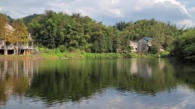 Small cottage houses on the shore of a lake surrounded by trees under a blue cloudy sky