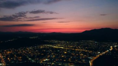 An aerial view of a city lit by yellow street lights, surrounded by hills under an orange-purple sky at sunset