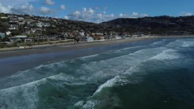 A Drone shot La Jolla Shores with wavy ocean and a view of coastal houses under a blue cloudy sky