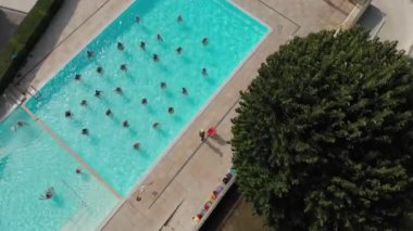 Yeşil tarlalar ve ağaçlarla çevrili bir havuzda su aerobiği yapan insanların kuş bakışı görüntüsü.