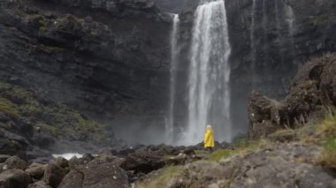 A blonde woman in a yellow raincoat in front of a waterfall in Faroe Islands, Denmark