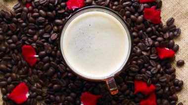 An overhead shot of a rotating cup of coffee on a pile of coffee beans with rose petals