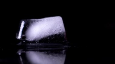 A close up time-lapse of an Ice cube melting into a puddle with a visible reflection isolated on a black background