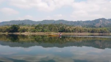 A man rowing a boat with speed across a calm, reflective lake surrounded by hills and forests under a bright sky on a sunny day
