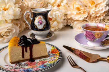 Masa örtüsü ve çiçeklerle çevrili kiraz reçelli bir dilim cheesecake.