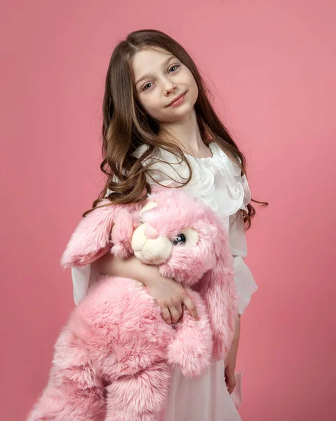 Küçük beyaz bir kızın pembe bir tavşanla poz verdiği dikey bir stüdyo resmi.