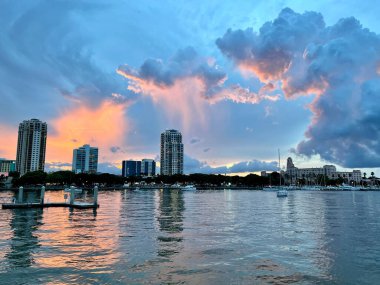 Mor bulutlu gün batımı gökyüzüne karşı su yansıtan kentsel yüksek kulelerin büyüleyici bir görüntüsü.