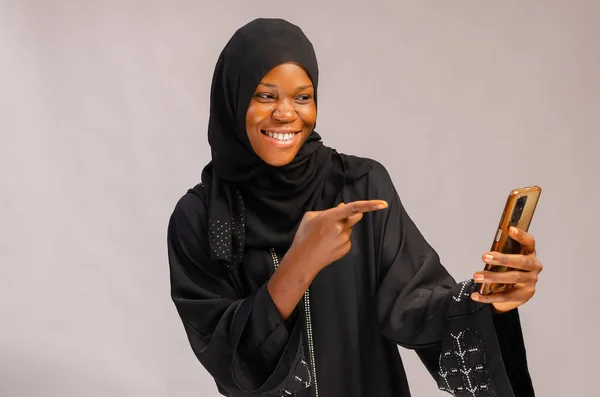 A beautiful African Muslim lady smiling and pointing to her phone with a light wall background
