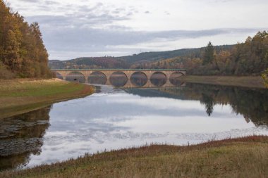 A beautiful view of a calm lake surrounded by green trees with a bridge in the background