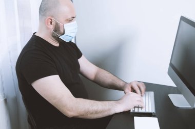 A closeup shot of a man wearing a black t-short and sitting next to a computer and a mask on his face