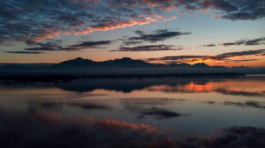 Göl ya da nehir manzaralı bir günbatımı gökyüzünü ve dağları yansıtıyor.