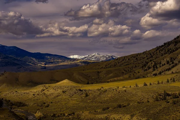 A VALLEY WITH ROLLING HILLS AND SNOW COVERED MOUNTAINS IN THE DISTANCE AND A CLOUDY SKY IN YELLOWSTONE NATIONAL PARK-