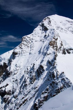 Mavi bulutlu gökyüzüne karşı karlı bir dağın dikey görüntüsü.