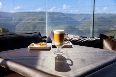 Kahve dükkanında kapuçino ve kiş kahvaltılı bir masa. Camdan güneş ışığı geliyor. Güneşli bir günde güzel bir dağ manzarası