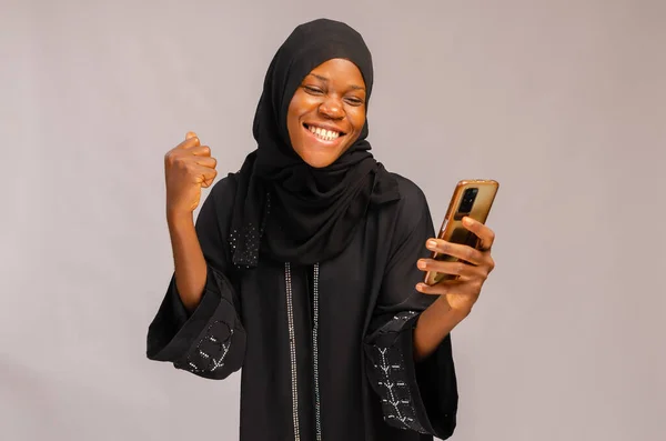 An African Muslim smiling excited lady using her phone and isolated on gray background