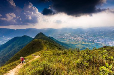 Dağların zirvelerinde bitkilerle ve bulutlu gökyüzüyle yürüyüş yapan insanlar.