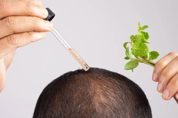 Man holding peppermint oil dropper applying on scalp to promote hair growth in grey background.