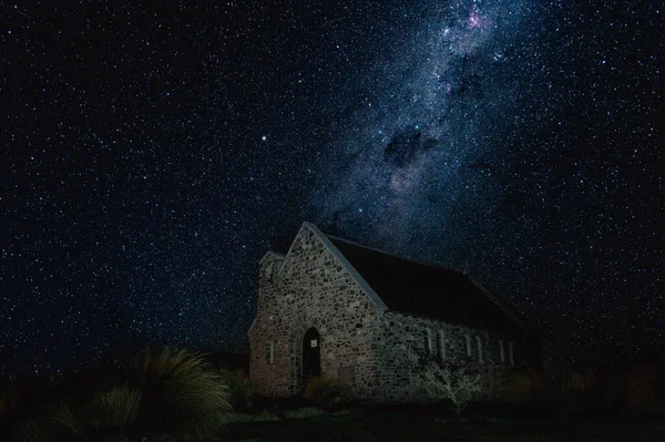 A view of a single-storey stone building in a rural area at night with the sky full of dense stars