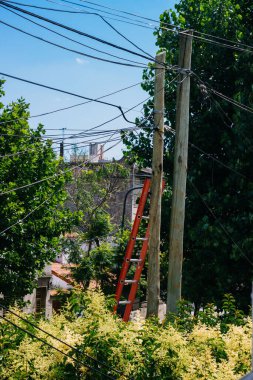 A vertical shot of a red ladder leaned on a light cables pillar outside on a sunny day