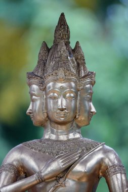 A vertical shot of a silver three-headed Buddha statue with greenery blurred in the background
