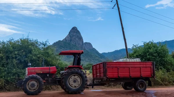 岩の多い山に対して農村部に駐車トロリー付きの赤いトラクター — ストック写真