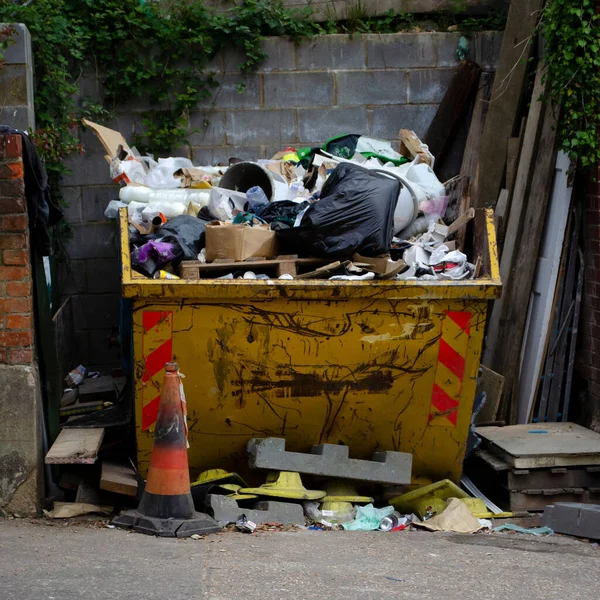 A full yellow rubbish skip full of trash from a building site. Wood and other building materials around it.  Ready to go to the dump.